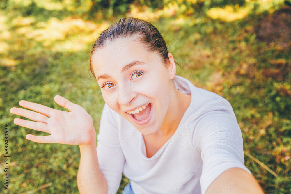 Young funny girl take selfie from hands with phone sitting on green grass park or garden background. Portrait of young attractive woman making selfie photo on smartphone in summer day. Young funny girl take selfie from hands with phone sitting on green grass park or garden background. Portrait of young attractive woman making selfie photo on smartphone in summer day.