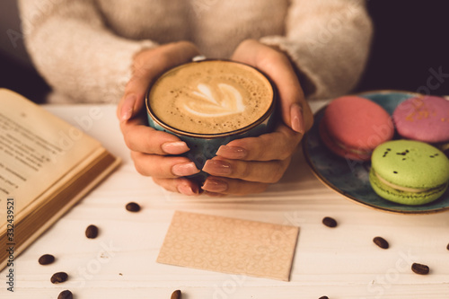 Beautiful female hands hold a blue cup with coffee. The girl is sitting at the table in the cafe. Nastoda open book, plate with colorful macaroons. photo