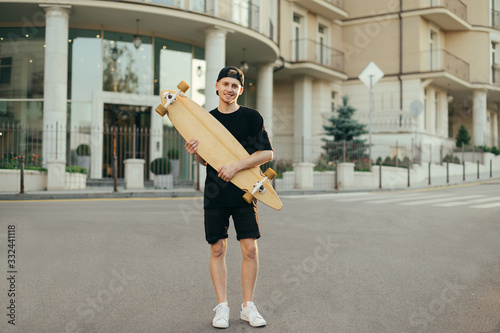 Full length portrait of teenager boy standing with longboard in hands outdoors, looking at camera and smiling. Young man in dark clothes posing at camera with skate in hands. Copy space