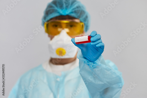 biochemist holding tube with red liquid to the camera in protactive suit with disposable medicl hat protactive mask and yellow glasses photo