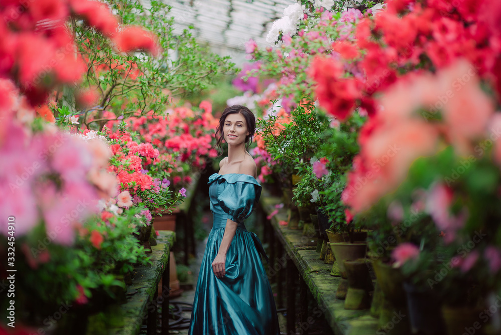 Beautiful romantic young woman in a greenhouse with azaleas. Art portrait of a girl wearing blue magnificent vintage dress.