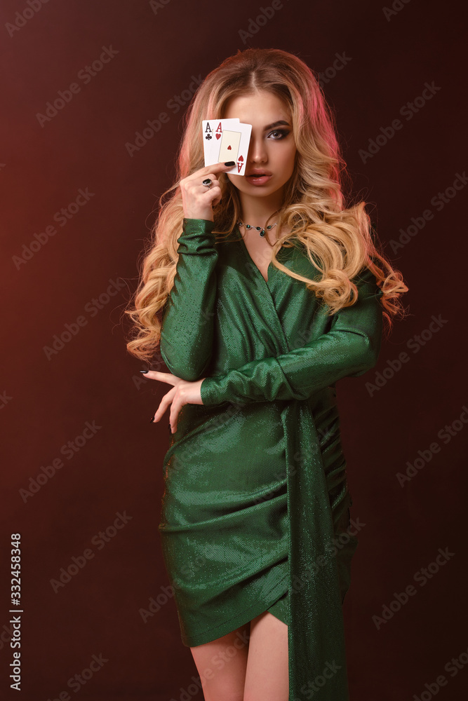Blonde curly woman in green dress and jewelry. She covered half of her face with two aces, posing on brown background. Poker, casino. Close-up