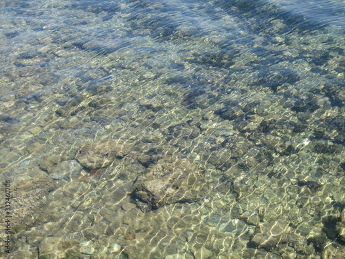 Through the clear water in the rays of the reflection of the sun, you can see the shallow sea floor of sand, pebbles and stones of a gray brown color.