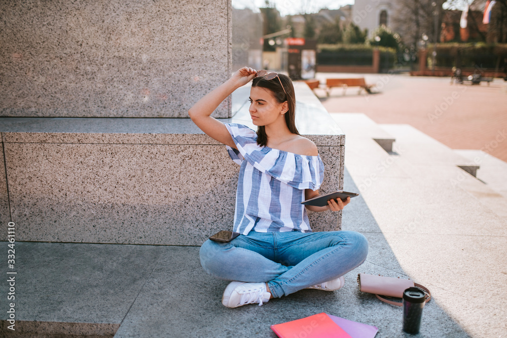 A young caucasian  beautiful girl sits next to a monument with a tablet in her hand