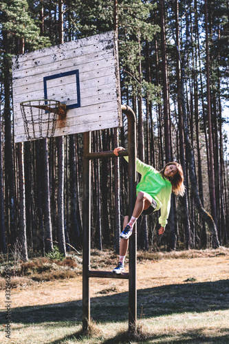 A brunette woman wearing a fluorescent green sweatshirt black shorts is dangling flirtatiously from a basketball stand.