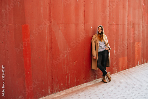 a young girl in a brown coat and glasses on the street, standing near a red wall.