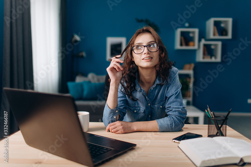 Happy beautiful woman in denim shirt playing with her curly hair while sitting at home with opened laptop. Dreaming girl in eyeglasses having break during working time.