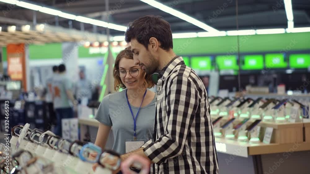 In the electronics store female consultant gives professional advice to a young man. He considers to buy a new smartphone and needs expert opinion. Store is modern, bright and has all the new devices In the electronics store female consultant gives professional advice to a young man. He considers to buy a new smartphone and needs expert opinion. Store is modern, bright and has all the new devices