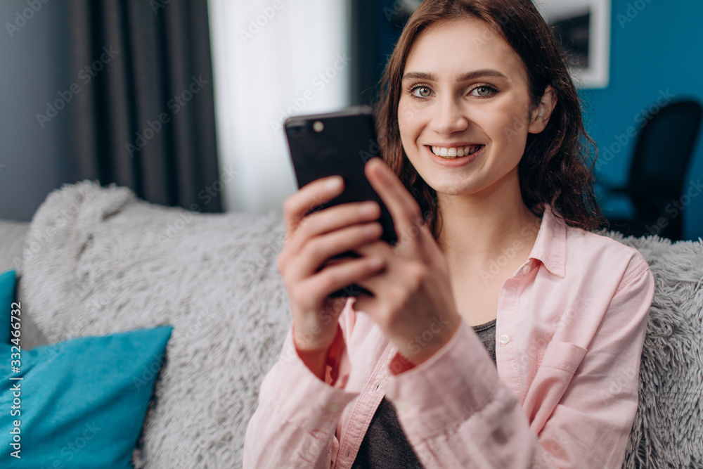 Cheerful young girl wearing home clothing, sitting on couch and using smartphone. Beautiful woman with curly hair smiling and looking at camera. Cheerful young girl wearing home clothing, sitting on couch and using smartphone. Beautiful woman with curly hair smiling and looking at camera.