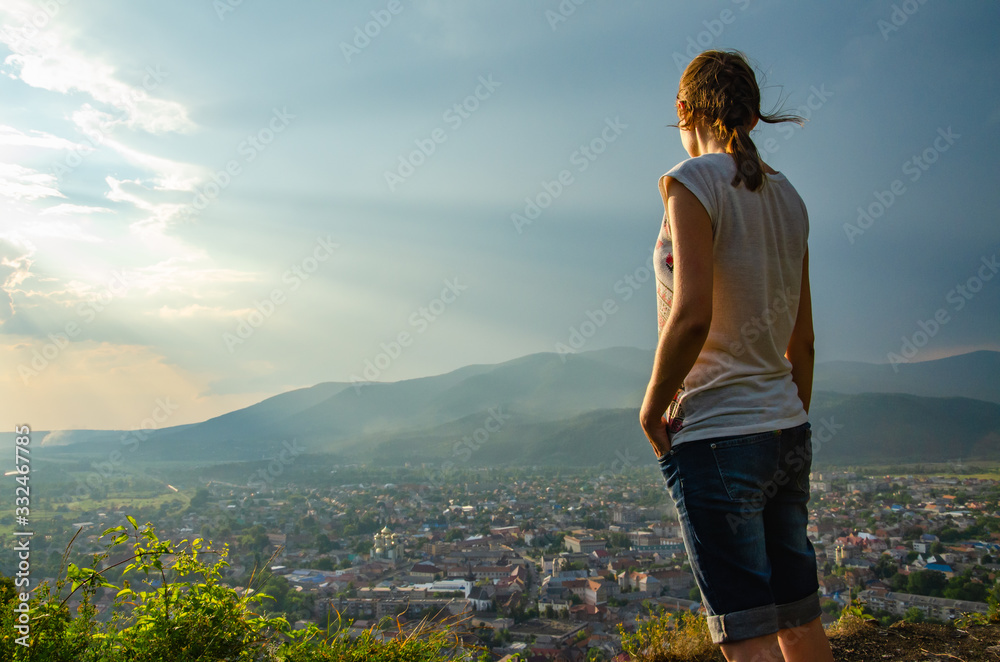 Naklejka premium Young woman standing on a hilltop against the backdrop of an epic sky above a small town in a valley among the mountains