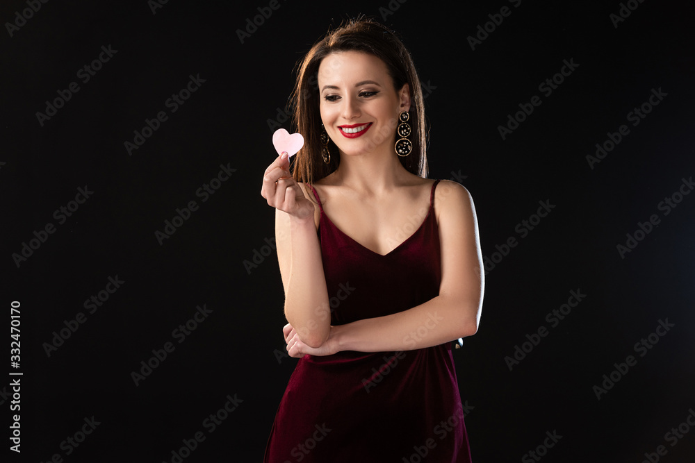 Fototapeta premium smiling woman in dress holding heart-shaped card isolated on black