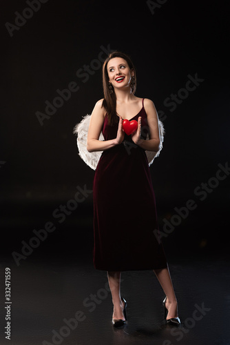 smiling woman in dress with wings holding heart-shaped model in 14 february on black background