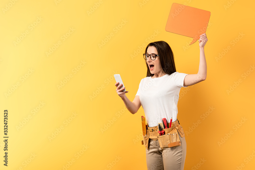 shocked repairwoman holding speech bubble and using smartphone on yellow background