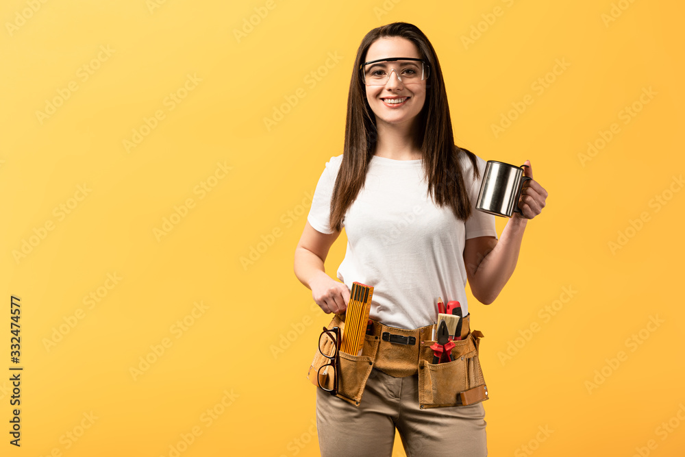 smiling handywoman holding metal cup and looking at camera isolated on yellow smiling handywoman holding metal cup and looking at camera isolated on yellow