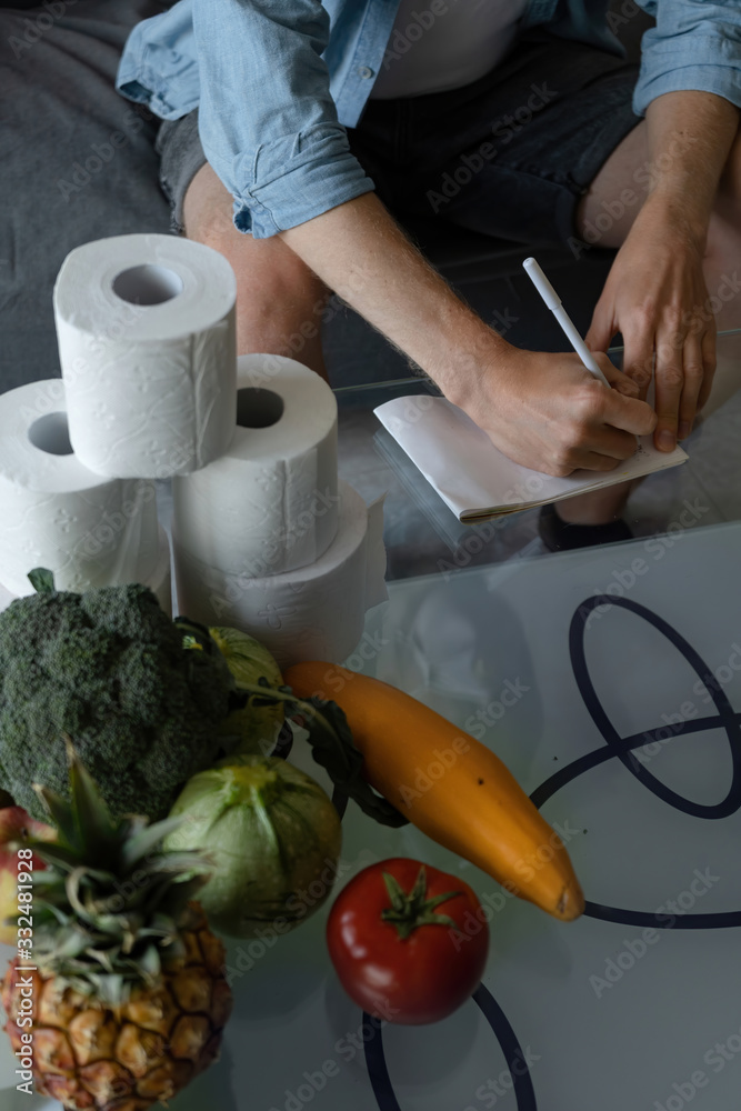 A guy sits in home quarantine with a supply of food for a month. Writes in a notebook the amount of different food meals. Covid-19 coronavirus pandemic in the world. Vegetables and fruits. A guy sits in home quarantine with a supply of food for a month. Writes in a notebook the amount of different food meals. Covid-19 coronavirus pandemic in the world. Vegetables and fruits.