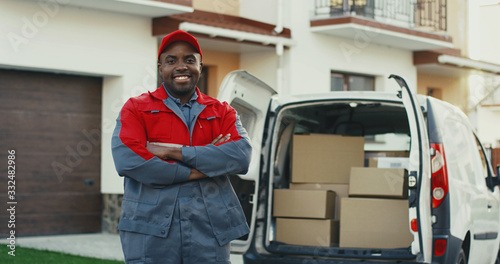 Joyful African American young deliveryman in the red uniform and cap standing at the white male van with boxes, crossing hands in front of him and smiling to the camera. Portrait shot. Outdoor.