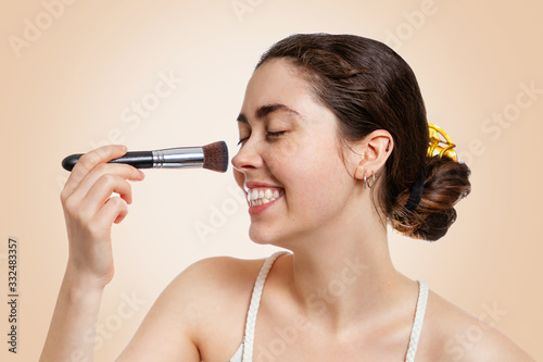 Portrait of a young happy woman with a smile applying makeup with a brush. Beige background. Concept of cosmetology, cosmetics and skin care