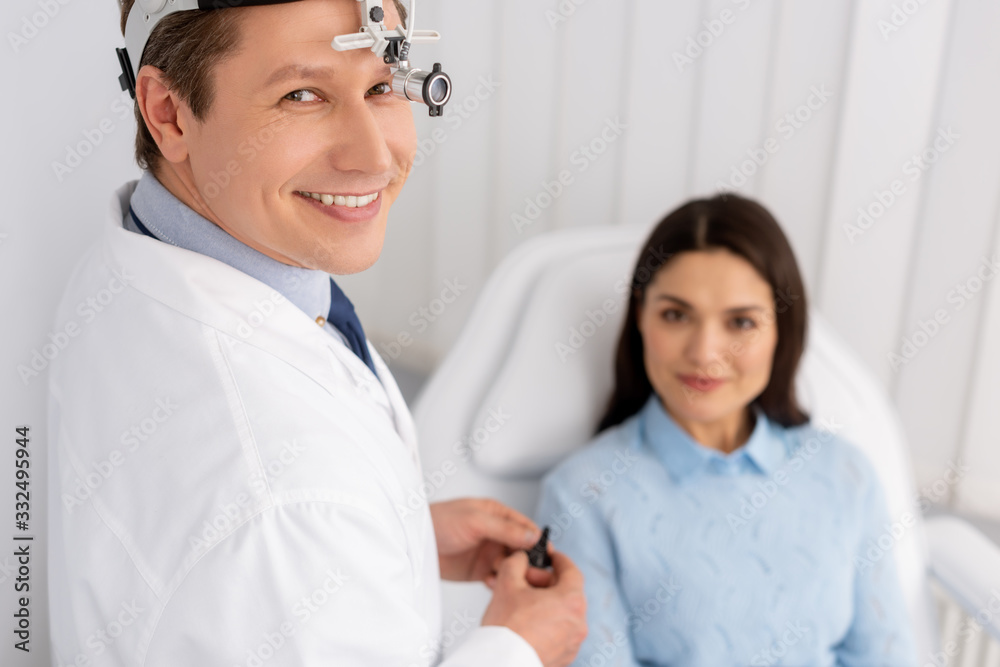 Fototapeta premium selective focus of smiling otolaryngologist in ent headlight holding ear speculum near smiling woman sitting in medical chair