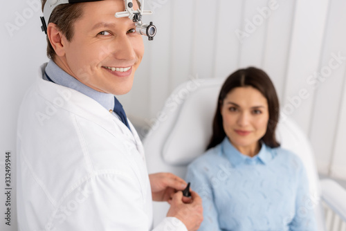 selective focus of smiling otolaryngologist in ent headlight holding ear speculum near smiling woman sitting in medical chair