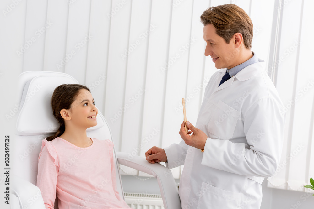 Fototapeta premium smiling otolaryngologist holding tongue depressor while standing near cheerful kid sitting in medical chair