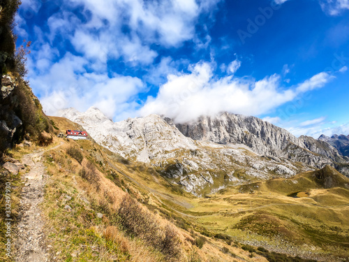 Pathway leading to an Alpine cottage in Italian Alps. Sharp slopes on both sides of the valley. Hard to reach mountain peaks. There are many mountain ranges in the back. Serenity and peace. Wandering