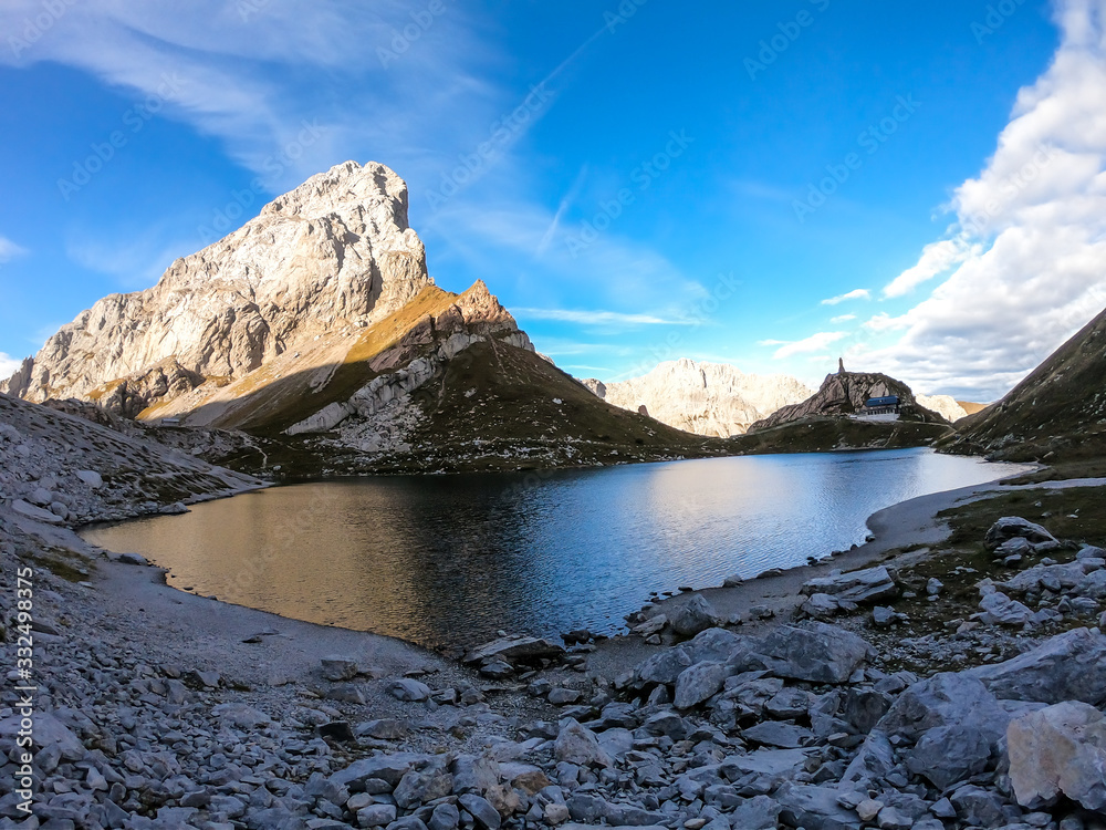 Obraz premium Panoramic shot of a reflection of an Alpine mountain in Wolayer Lake Austria. Completely still surface of the lake. Mountain is catching the first sunbeams. Alpine cottage on the side. High altitude