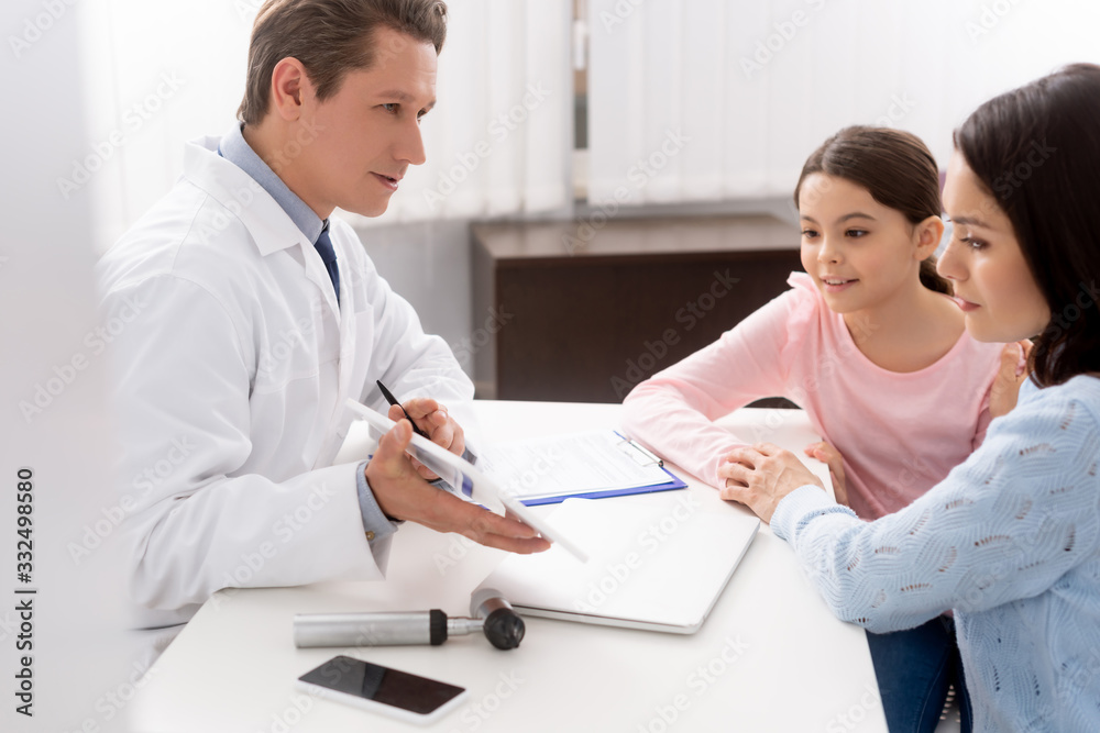 selective focus of otolaryngologist showing digital table to mother and daughter during consultation selective focus of otolaryngologist showing digital table to mother and daughter during consultation