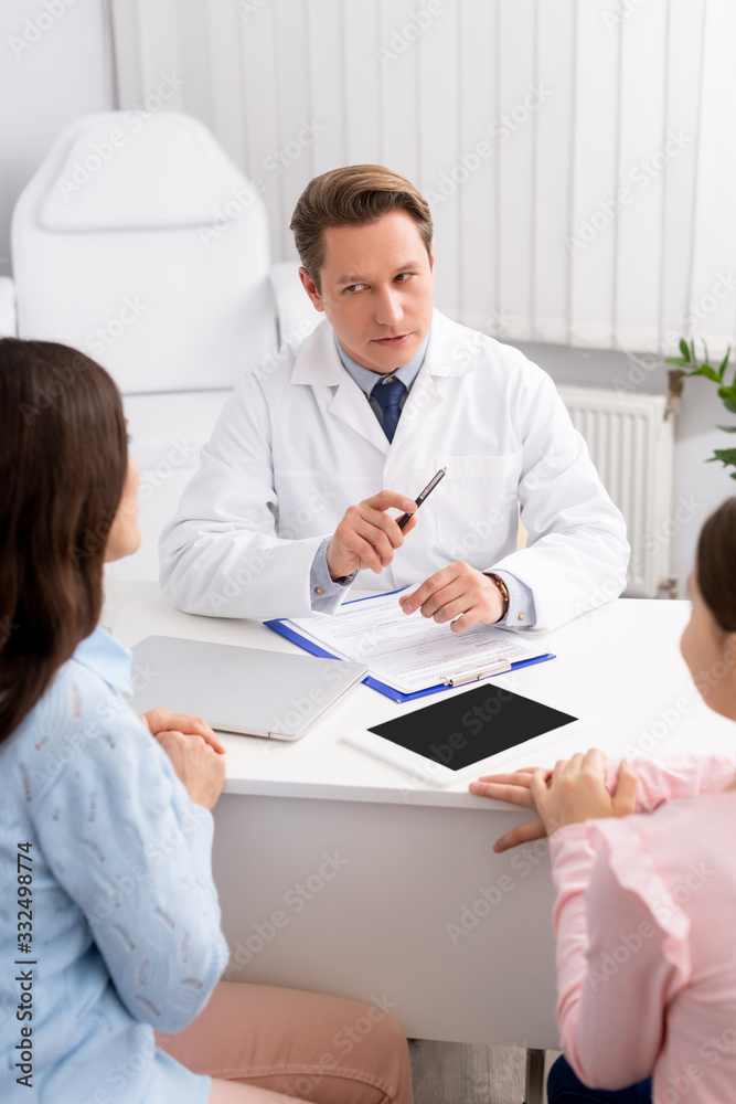 Fototapeta premium serious otolaryngologist talking to mother and daughter near digital tablet with blank screen on desk