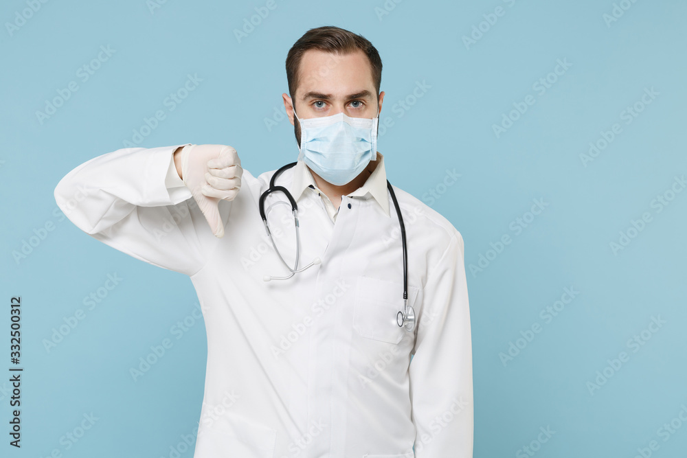Male doctor man in white medical gown sterile face mask gloves isolated on blue wall background. Epidemic pandemic spreading coronavirus 2019-ncov sars covid-19 flu virus concept. Showing thumb down. Male doctor man in white medical gown sterile face mask gloves isolated on blue wall background. Epidemic pandemic spreading coronavirus 2019-ncov sars covid-19 flu virus concept. Showing thumb down.