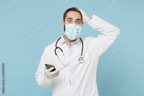 Male on-line doctor man in medical gown sterile face mask gloves isolated on blue background. Epidemic pandemic coronavirus 2019-ncov sars covid-19 flu virus concept Use mobile phone put hand on head.