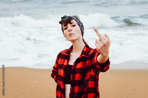 Arrogant defiant young female with piercing and cigarette wearing unbuttoned shirt showing middle finger while standing on sandy beach with sea waves in background
