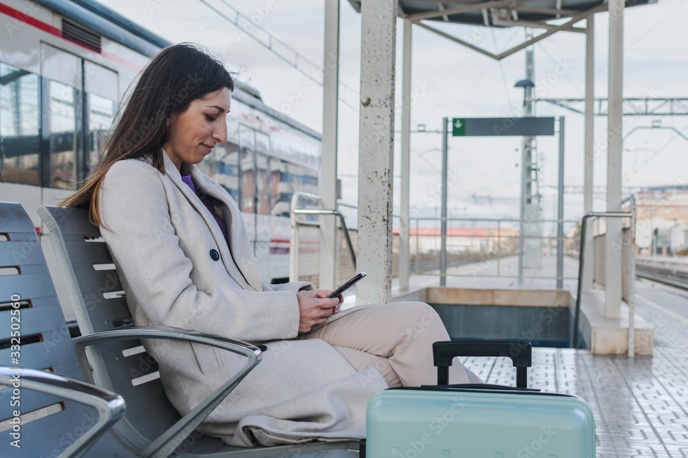 Side view of modern female traveler with suitcase sitting on bench on platform of railway station and using smartphone while waiting for train Side view of modern female traveler with suitcase sitting on bench on platform of railway station and using smartphone while waiting for train