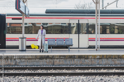 Young female traveler in stylish outfit standing with yellow balloons in hand and suitcase on platform of railway station