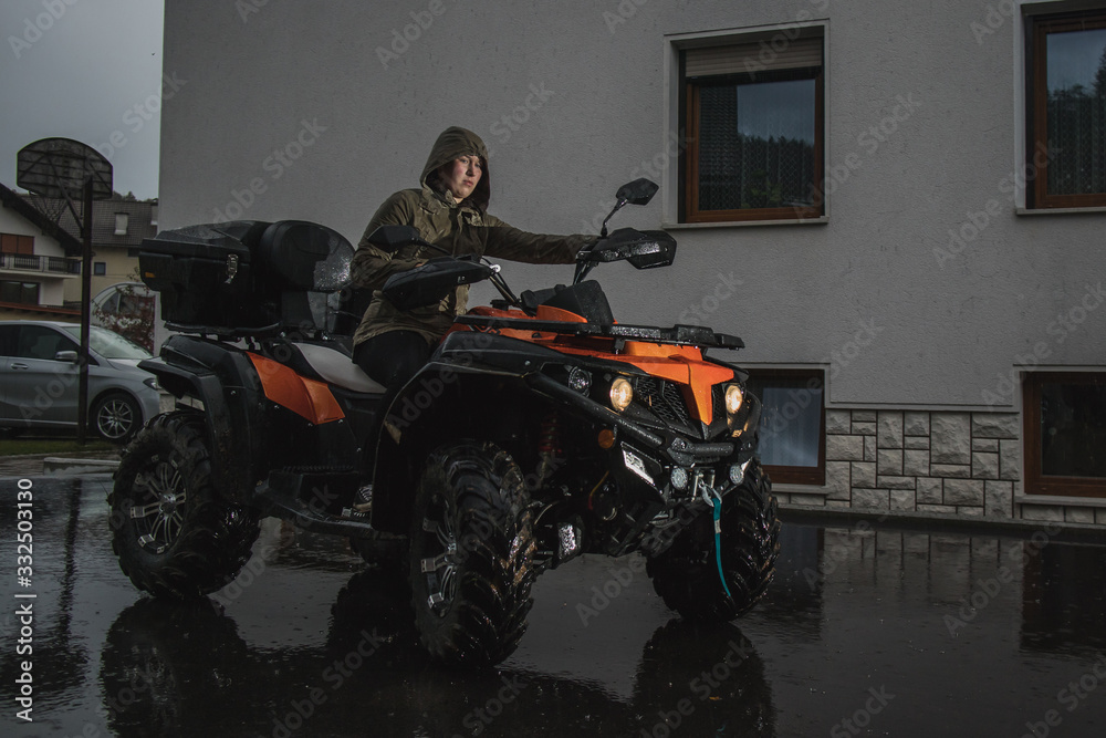 Young caucasian woman is manouvering an orange quad or ATV vehicle while it is raining outside of the house.