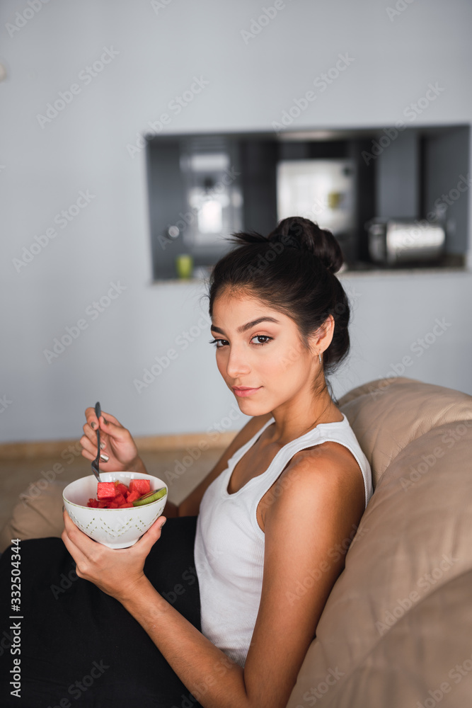 Side view beautiful brunette Hispanic woman holding fork and bowl of kiwi and watermelon and sitting on settee Side view beautiful brunette Hispanic woman holding fork and bowl of kiwi and watermelon and sitting on settee