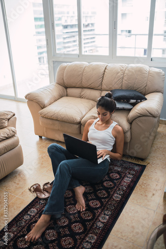From above beautiful brunette Hispanic woman typing on laptop and sitting on floor on little carpet near settee and window