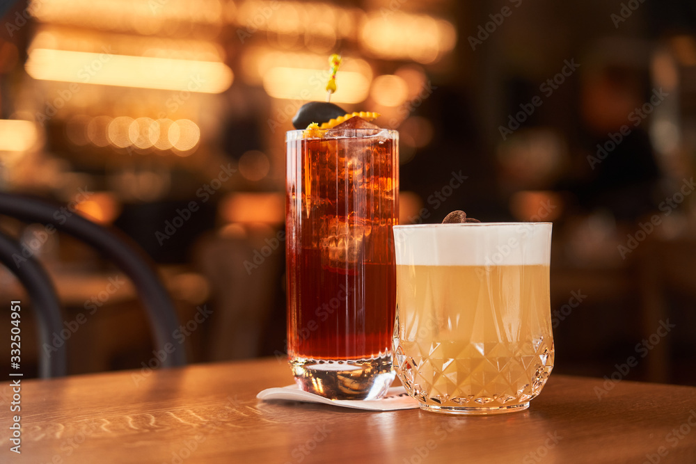 Cocktail with ice cubes decorated with stick with black olive and Whiskey Sour with lemon juice cocktail on wooden counter