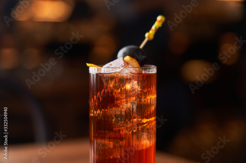 Highball glass with red alcohol cocktail with ice cubes decorated with stick with black olive on wooden counter photo