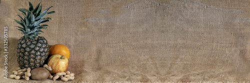 Horizontal banner. Pineapple orange apple and peanuts on raw yuta canvas background.