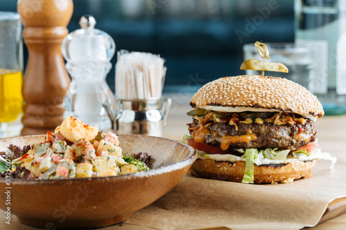 Big hamburger on paper with cutlet cheese and vegetables served with bowl of colorful salad in modern restaurant