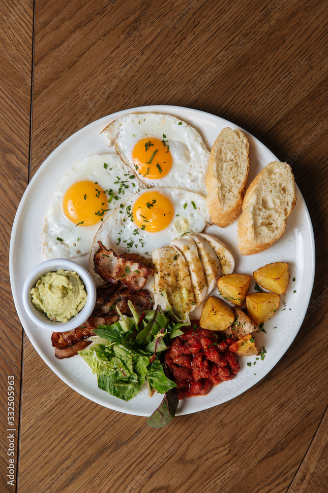 Top view of served English breakfast with fried eggs and bacon with toasts and vegetables on wooden table Top view of served English breakfast with fried eggs and bacon with toasts and vegetables on wooden table