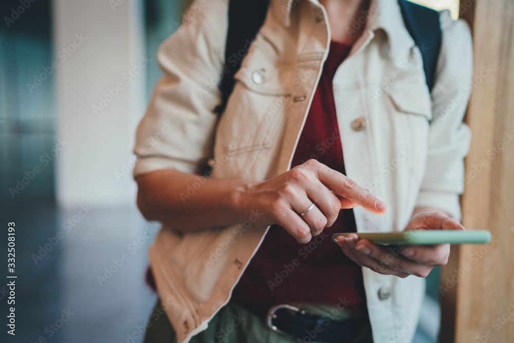 Fototapeta premium Cropped image of young hipster student girl in educational building typing a message on the touch screen or playing an online game on her mobile phone during lunch break