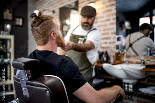 Barber trimming beard of redhead man sitting in barbershop photo