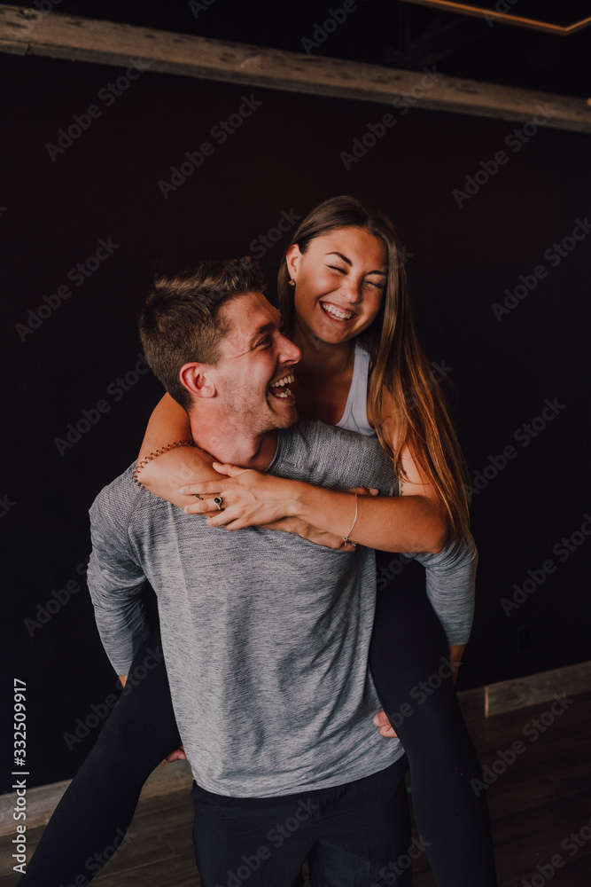 Excited adult man in sportswear piggybacking slender young woman while standing against black wall in contemporary gym Excited adult man in sportswear piggybacking slender young woman while standing against black wall in contemporary gym