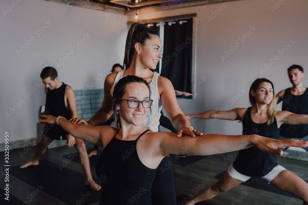 Smiling diligent female instructor in sportswear teaching warrior pose two group of sporty people doing exercise standing on sports mats in modern workout room Smiling diligent female instructor in sportswear teaching warrior pose two group of sporty people doing exercise standing on sports mats in modern workout room
