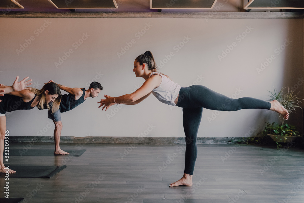 Slim barefoot female instructor in sportswear standing in warrior pose three and controlling group of sporty people concentrating and doing same exercise standing on sports mats in modern workout room