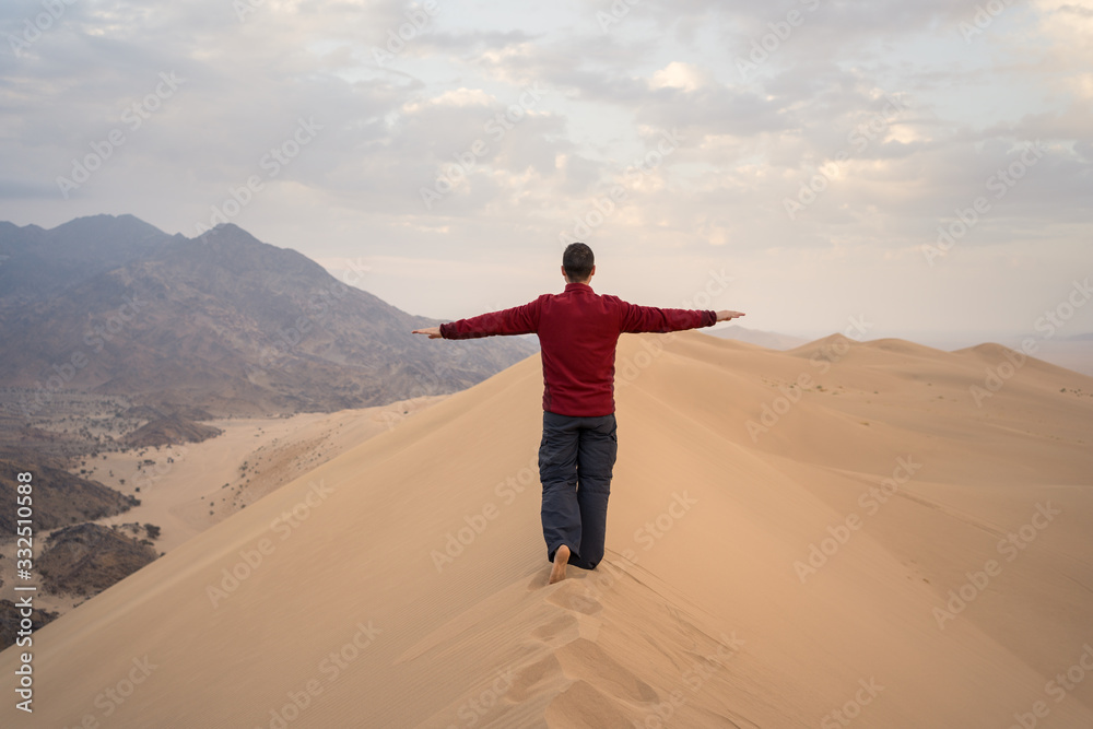 Back view of unrecognizable happy man with arms raised in casual clothes standing on dune during tour to desert in Saudi Arabia Back view of unrecognizable happy man with arms raised in casual clothes standing on dune during tour to desert in Saudi Arabia