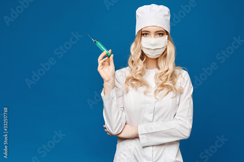 Pretty female doctor wearing white medical robe holding syringe at blue background, isolated. Healthcare and pharmacology concept. Young woman in medical uniform and protective mask prepares a syringe