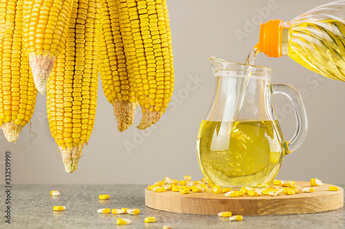 Pouring Corn oil in glass bottle with dried corn groats and kernels on rustic backdrop. Cooking oil composition photo