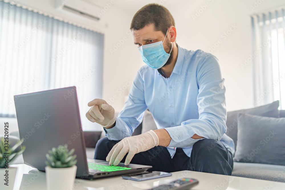 Young adult one man caucasian wearing gloves on hands and protective mask on face while working from home sitting by the table laptop preventing virus spread in epidemic quarantine health prevention